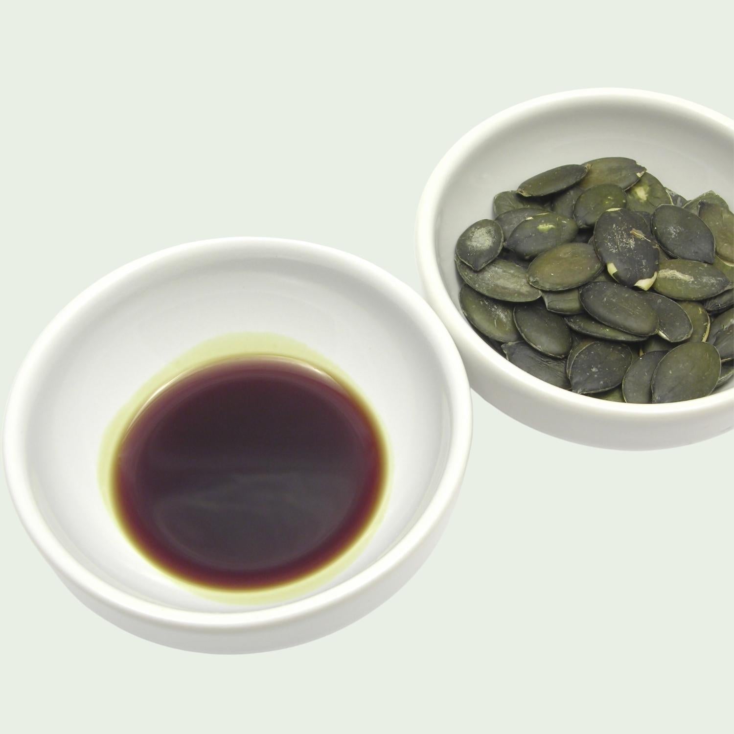 Two white bowls, one with dark green Styrian pumpkin seed oil and the other with Australian pumpkin seeds, on a light gray background.
