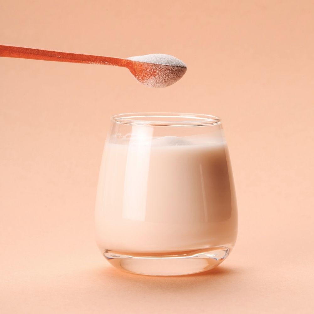 Glass of Collagen peptides with a spoonful of easily absorbed neutral tasting Collagen powder above it on a peach background