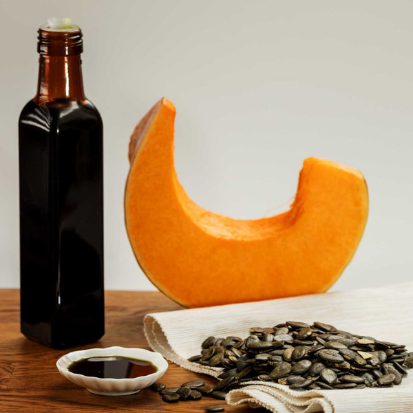 Bottle of dark Australian pumpkin seed oil, wedge of orange Styrian pumpkin, and bowl of green pumpkin seeds on a wooden surface.